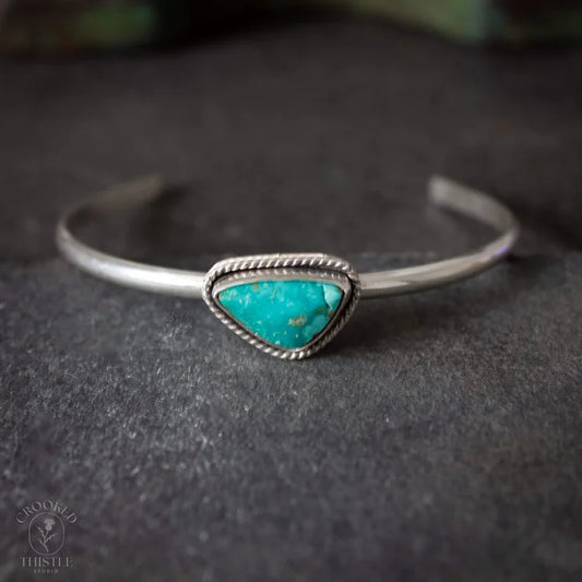 A sterling silver cuff bracelet featuring a triangular turquoise stone centerpiece, resting on a dark textured surface. Crooked Thistle Carico Lake Turquoise Bracelet