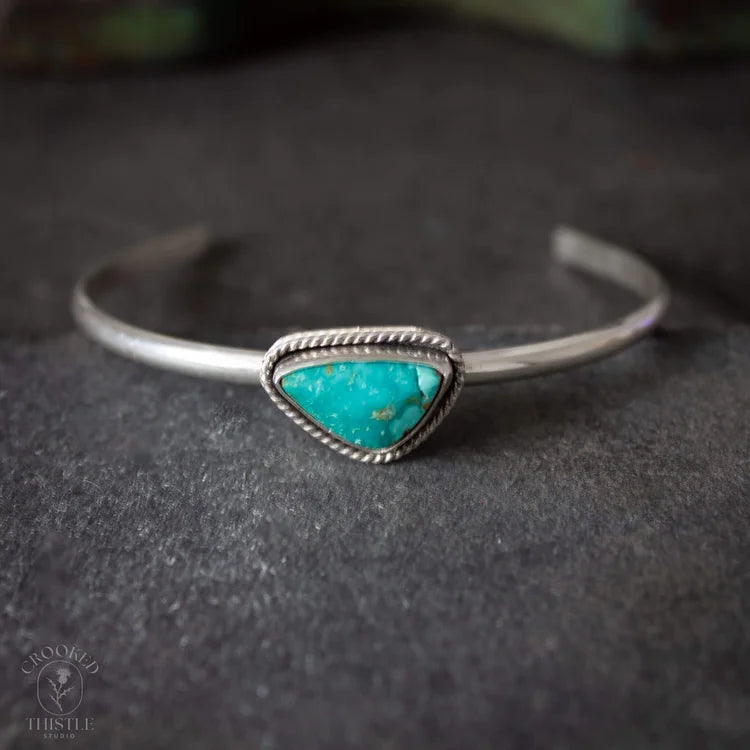 A sterling silver cuff bracelet featuring a triangular turquoise stone centerpiece, resting on a dark textured surface.  Crooked Thistle Carico Lake Turquoise Bracelet